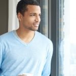 A young guy staring out a window holding coffee and smiling