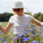 A woman standing on a field of violet flowers. She looks happy and is waring a hat and sunglasses.