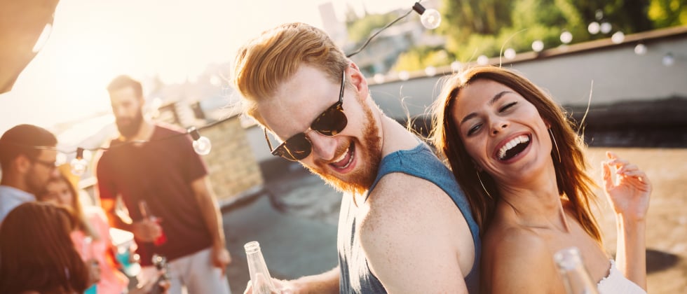 A man and a woman laughing and dancing back-to-back at a rooftop party.
