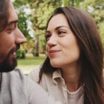 A man and a woman sitting next to each other in nature. She is looking at him happily and showing a love language.