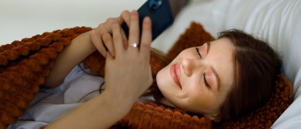 A woman laying in bed with her phone, showing signs she wants you to ask her out.