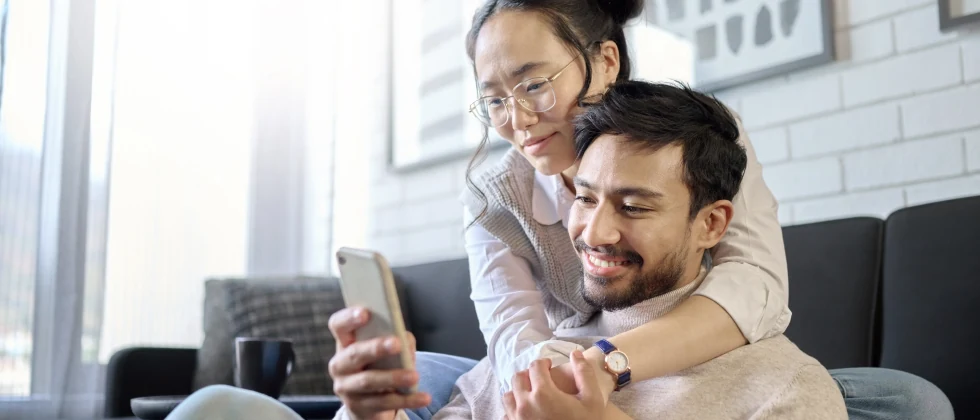 A woman with her arms around a man's neck, looking at a phone.
