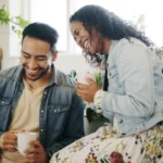 A couple sitting on the kitchen corner holding cups and laughing.